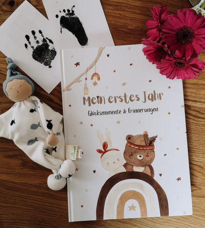 Eulentaler Babyalbum „Mein erstes Jahr – Glücksmomente & Erinnerungen“ mit Waldtiermotiven neben schwarzen Baby-Hand- und Fußabdrücken, Schmusetuch und pinken Blumen.