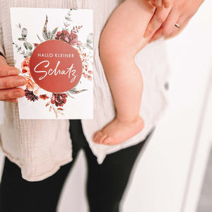 Frau hält Meilensteinkarte "Hallo kleiner Schatz" während sie ein Baby auf dem Arm trägt.