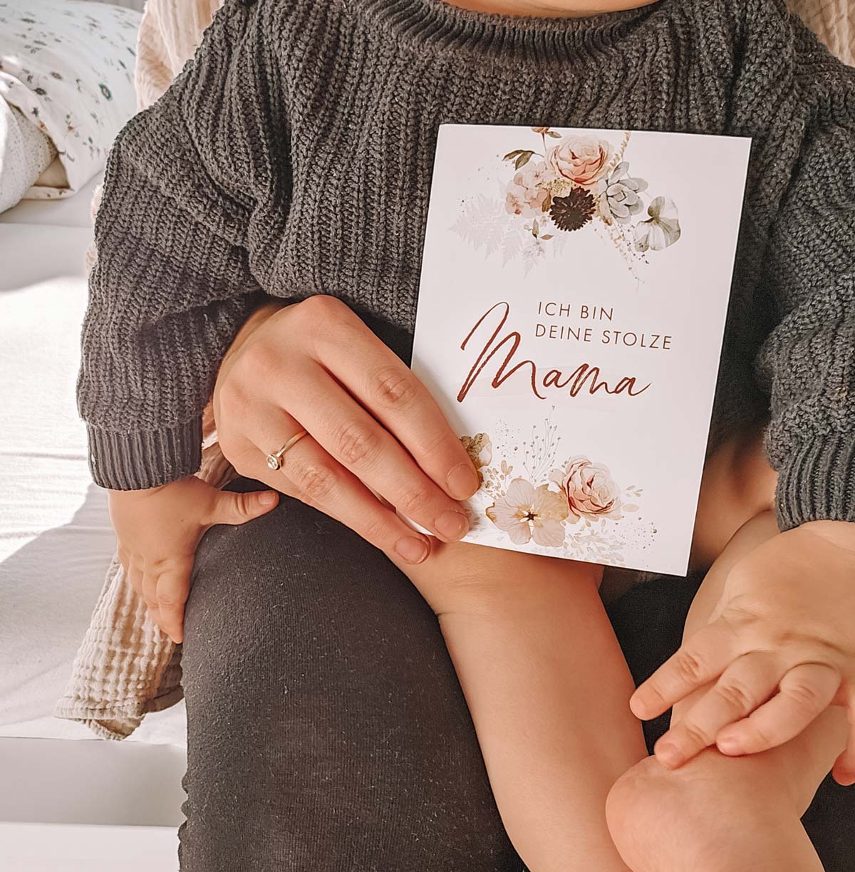 Mutter hält Meilensteinkarte "Ich bin deine stolze Mama" mit Baby auf dem Schoß.