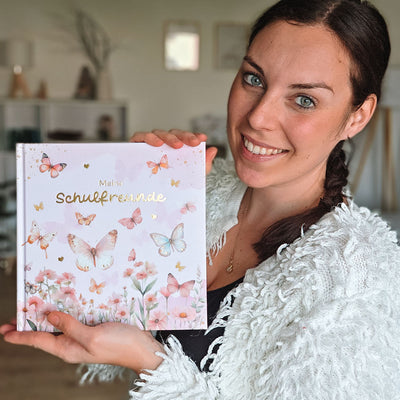 Frau hält lächelnd das Eulentaler Schulfreundebuch „Meine Schulfreunde“ mit Schmetterlingen, Blumen und goldgeprägtem Titel in die Kamera.
