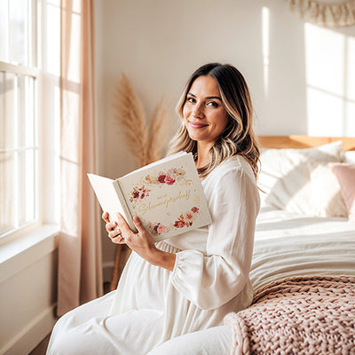 Schwangere Frau sitzt auf dem Bett und hält das Eulentaler Schwangerschaftstagebuch „Meine Schwangerschaft“ mit floralem Cover und Goldprägung. 
