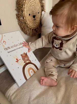 Baby sitzt neben dem Eulentaler Babyalbum „Mein erstes Jahr“ mit Waldtiermotiven und Regenbogen auf dem Cover.
