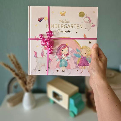 Hand hält das Eulentaler Kindergarten-Freundebuch mit Einhorn- und Feenmotiv, verziert mit einem pinken Geschenkband.
