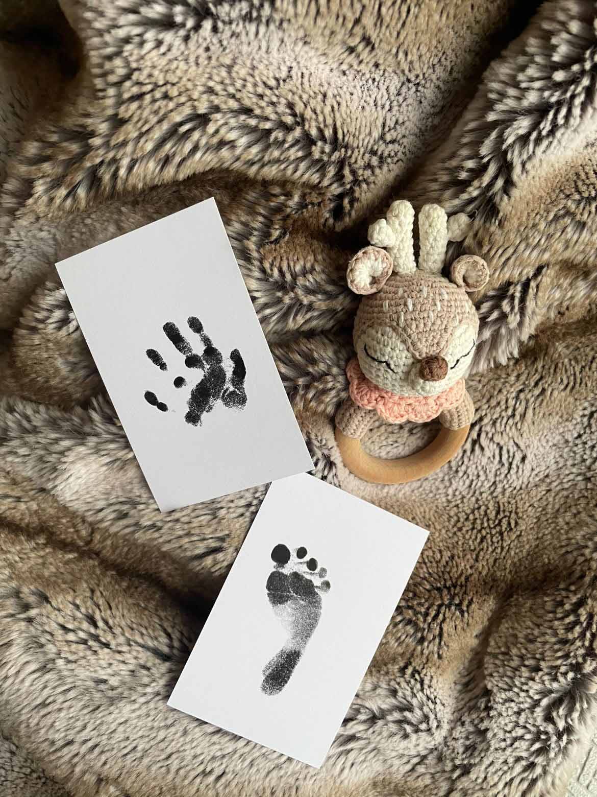 Schwarzer Hand- und Fußabdruck eines Babys auf weißen Karten neben einer gehäkelten Reh-Rassel auf einer weichen Felldecke.

