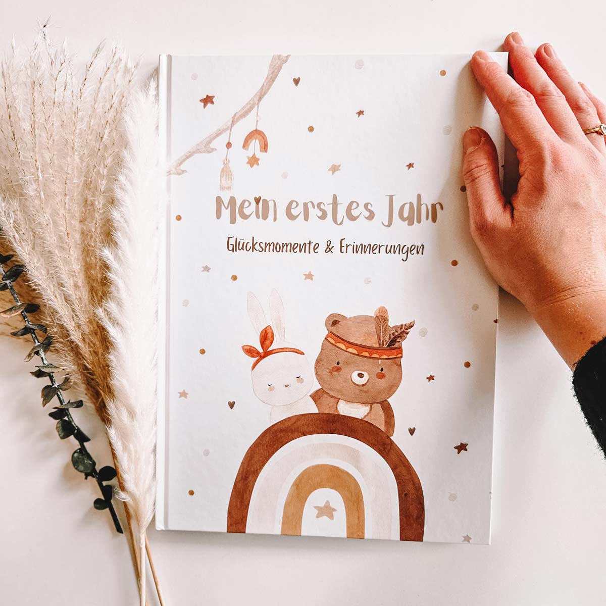 Hand hält ein Babytagebuch 'Mein erstes Jahr' mit niedlichen Tierfiguren und Regenbogen auf dem Cover, neben Pampasgras.