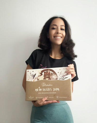 Frau hält lächelnd die Eulentaler Erinnerungsbox „Mein erstes Jahr“ mit Babyalbum, Abdruckset und Meilensteinkarten in den Händen.
