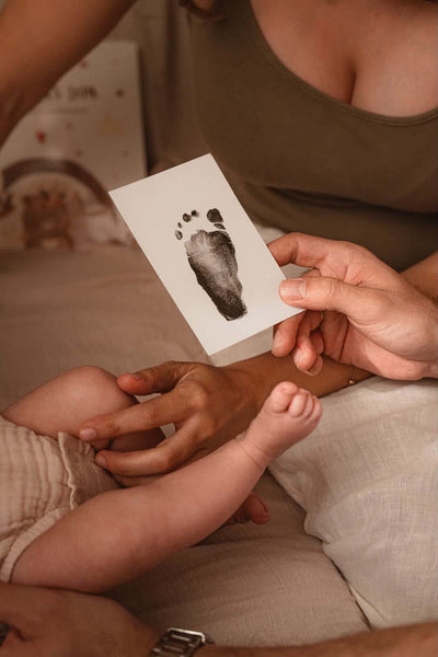 Eltern halten neben ihrem Baby eine Karte mit schwarzem Fußabdruck als Erinnerung an die ersten Lebensmonate.
