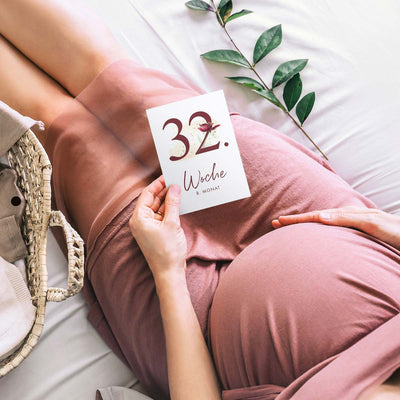 Schwangere Frau hält Meilensteinkarte "32. Woche" auf ihrem Babybauch liegend.