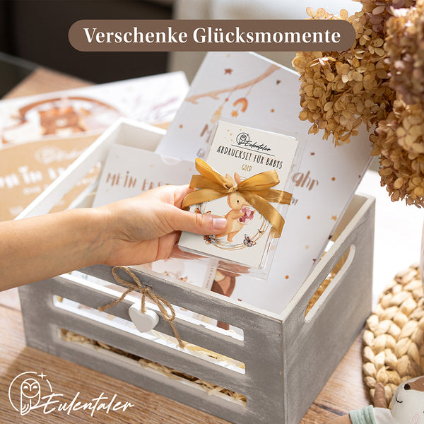 Hand hält das Eulentaler Abdruckset für Babys in Gold mit Geschenkband über einer dekorativen Geschenkbox.
