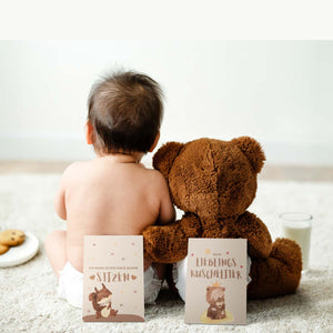Baby sitzt mit einem Teddybär auf dem Boden, beide halten Meilensteinkarten aus dem Set "Mein Erstes Jahr".