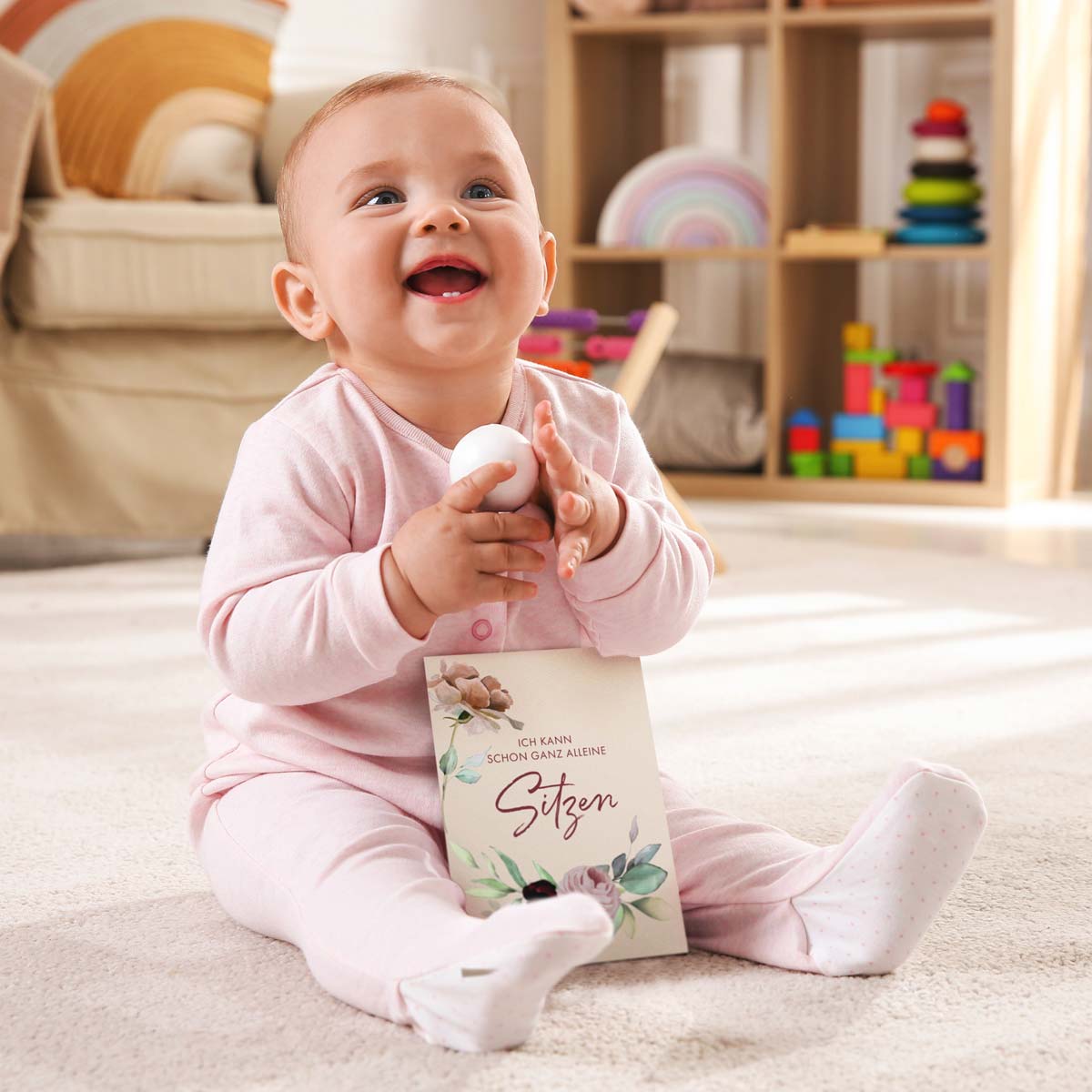 Glückliches Baby sitzt alleine mit einer Meilensteinkarte in der Hand auf einem Teppich im Kinderzimmer.