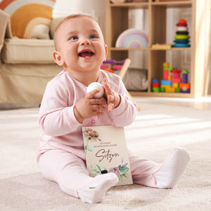 Glückliches Baby sitzt alleine mit einer Meilensteinkarte in der Hand auf einem Teppich im Kinderzimmer.