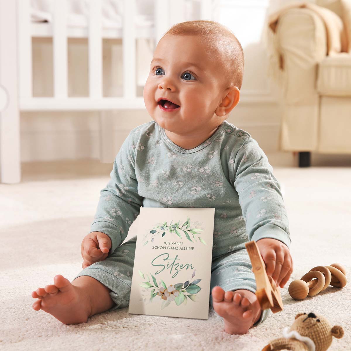 Lächelndes Baby sitzt auf dem Boden und hält eine Meilensteinkarte mit der Aufschrift "Ich kann schon ganz alleine sitzen".