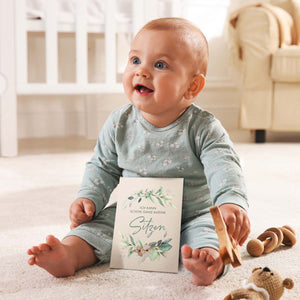 Lächelndes Baby sitzt auf dem Boden und hält eine Meilensteinkarte mit der Aufschrift "Ich kann schon ganz alleine sitzen".