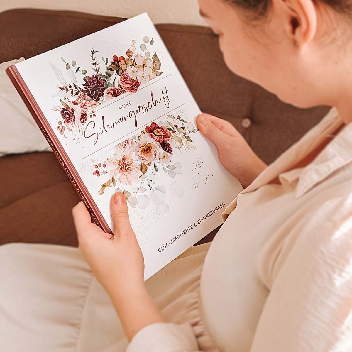 Schwangere Frau betrachtet liebevoll ein Schwangerschaftstagebuch mit floralen Verzierungen auf dem Cover.