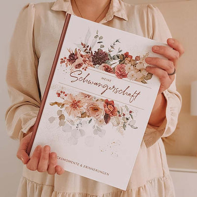 Frau hält ein schön gestaltetes Schwangerschaftstagebuch mit floraler Verzierung in den Händen.
