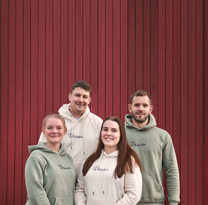 Vier Mitglieder des Eulentaler-Teams stehen lächelnd in Eulentaler-Hoodies vor einer roten Holzwand.
