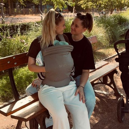 Zwei Frauen vom Instagram-Account @accouple sitzen mit einem Baby in der Trage auf einer Parkbank neben einem Kinderwagen und schauen sich liebevoll an.
